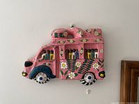 Front view of hand-painted papier-mâché bus showing pink body, floral decorations, small painted figures, and black wheels.