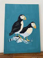 Full view of the painting showing two puffins on a teal background, standing on rocks.