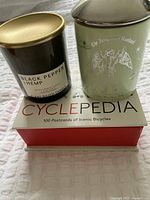 Photo of two soy candles and a Cyclepedia box with 100 postcards of iconic bicycles on a white textured surface.