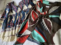 Photo showing two blouses: a multicolor geometric and leaf pattern blouse with rich colors including red, blue, black, and cream, alongside a blouse with a large abstract leaf print in brown, gray, red, and teal.