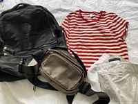 Photo showing black Lululemon backpack, beige fanny pack, red and white striped short-sleeve shirt, white shorts arranged on a white surface.