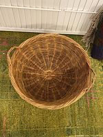 Top-down view of a large round wicker basket showing the intricate weaving pattern and two side handles. Basket rests on a green patterned carpet.
