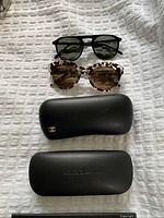Two pairs of sunglasses with three eyewear cases laid out on a white textured surface.