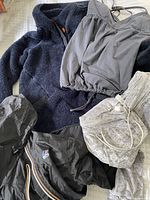 Navy fleece jacket, gray velvet leggings, gray strappy top, and K-Way black jacket with hood and multicolor zipper trim laid out on a bed