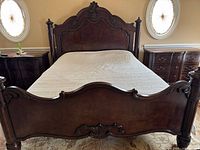 California King Bed with visible ornate carved headboard and footboard, two matched nightstands, and dresser in the background with similar style and finish.