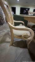 Side view of a light wood dining chair showing curved armrests with scroll design, vertical slat back, and beige fabric seat cushion.