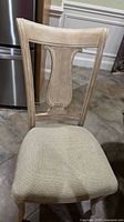 Front view of a wooden dining chair with a carved lyre back and beige upholstered seat, placed on a tiled floor.