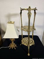 Brass table lamp next to ornate brass two-tier plant stand on black cloth