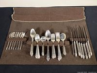 Cutlery pieces laid out on a brown cloth showing knives, soup spoons, forks, and spoons with ornate King Cedric pattern handles.