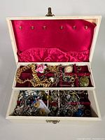 Open white jewelry box showing pink satin and red velvet compartments filled with assorted gold-tone and silver-tone costume jewelry including chains, brooches, and earrings.
