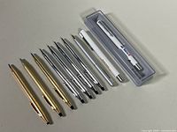 Seven ballpoint pens laid out including four silver Cross pens, three gold Cross pens, and two white promotional pens plus one Parker pen in plastic case