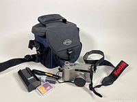 Photo showing the Kodak DC4800 digital camera alongside a lens cover, Kodak battery charger, AA battery, SanDisk 48MB memory card, and the Optex branded black and blue camera case with shoulder strap.