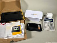 Photo showing all three main items in the lot: Kodak digital picture frame in original box, Humanware magnifier with box and travel sleeve, Sharp calculator with printing paper roll.
