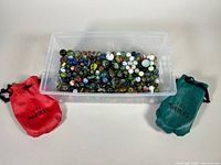 Plastic bin filled with assorted clear and opaque marbles with two fabric pouches, one red and one teal, each labeled '101 Marbles'.