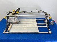 Full view of Mastercraft powered wet tile cutter on protective blue blanket, showing entire metal frame and cutting platform with circular blade on sliding mechanism.