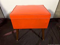 Front view of the orange vinyl upholstered sewing stool with wooden legs.