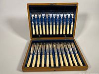 The full set of twelve fish forks and twelve matching fish cutters displayed inside the wooden box with the box lid open revealing the blue velvet lining.