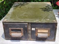 Front view of the 1950s metal filing cabinet showing both drawers with label holders and metal handles. Olive green top and black drawer fronts with surface wear and scratches visible.