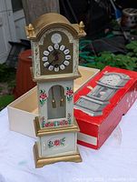 Miniature grandfather clock standing next to its original box, showing front view with decorated clock face, pendulum, and floral details.