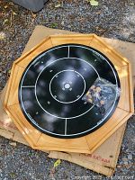 Photo of the octagonal crokinole board on cardboard with a bag of wooden game pieces placed on top, showing the playing surface and wood frame detail.