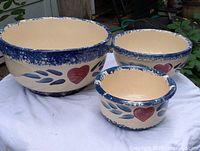 Three heart decorated ceramic mixing bowls shown from side on white cloth, with textured blue rims and red hearts with blue leaves on cream base.