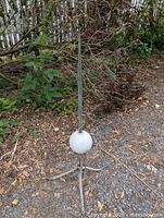 Full view of the antique lightning rod standing outdoors on gravel, showing the slender metal rod with a white glass ball near its base and three-legged mounting bracket.