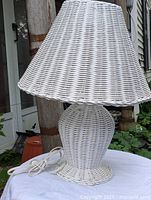 Front view of antique style white wicker table lamp showing the lamp base, conical lampshade, white power cord coiled on the base, photographed outdoors.