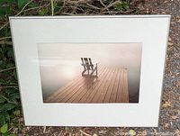 Photo of framed artwork showing an Adirondack chair on a wooden dock on a misty lake with sunrise light, housed in a white frame with white matting.