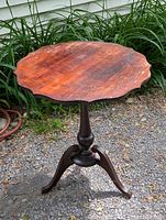 View of entire wooden pedestal side table showing scalloped pie crust edge top and tripod base in outdoor setting with gravel underfoot and greenery in background.