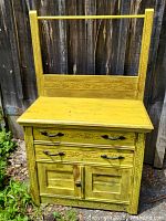 Front view of a yellow-painted oak washstand with carved floral details, two drawers and a cabinet below, standing outdoors against a wooden fence.