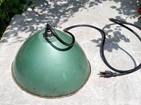 Photo of top and side view of green metal barn lamp with black electrical cord and plug shown, resting on a concrete surface with foliage shadows in background.