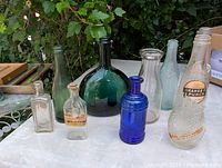 Overview of all 9 vintage glass bottles displayed on white table with greenery background.