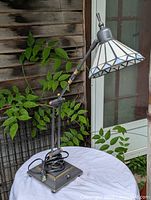 Full view of the desk lamp on a white cloth-covered round table outdoors, showing the stained glass-style shade, moving arm, and weighted base.