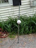 Full view of tall metal lightning rod with milk glass ball mounted on metal tripod supports in garden.