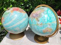 Two desktop globes on gold-tone metal bases placed outdoors on a table with garden plants in the background. Each globe shows detailed world maps with pastel blue oceans.