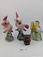 Five bird figurines: two pink cockatiels (one with wing repair), a brown ceramic owl, and a pair of porcelain birds in yellow, purple, and green base.