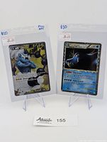 Two Pokemon cards propped on clear stands, showing front artwork, pricing tags, handwritten notes marking prices $20 and $30