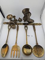 Photo showing the brass utensil rack with four hanging brass utensils and several small brass trinkets including a decorative brass boot and a small perforated bell.