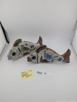 Two ceramic fish sculptures side by side showing bird and floral motifs with earthy colors, one fish's tail visibly repaired.