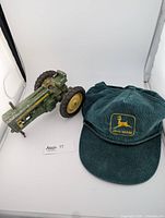 Photo showing vintage green and yellow diecast John Deere tractor with some paint wear alongside a dark green corduroy John Deere cap with embroidered logo patch and a black decorative cord above brim.