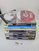 Stack of Xbox and Xbox 360 games including disc-only and full cases visible, titles partially readable showing variety of games.