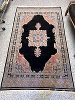 Full view of the Persian style wool rug showing overall design, coloration with black central field and pink, blue, cream borders, and fringes.