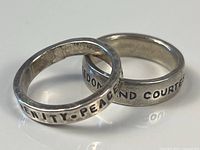 Two sterling silver rings intertwined on a white reflective surface showing inscribed inspirational words.