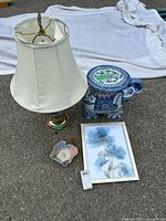 Photo showing the lamp with torn cream shade, elephant mini stand, blue floral framed print, and small ceramic bowl on a paved surface with white fabric background.