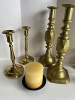 Photo showing all four brass candlesticks and a small beige pillar candle on a black base.