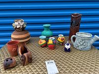 Lot of various decorative vases, planters and wood decorations arranged on a quilted golden surface, taken from a front view. Includes wooden vase with floral detail, turquoise pottery vase, small colorful ceramic vases, a gray metal watering can planter, and other small decorative items.