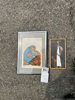 Both framed items displayed on concrete: left silver framed signed print with mother and children artwork, right wood framed tile with stylized figure and animal.