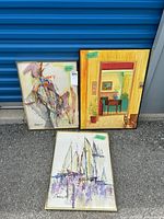 Three framed watercolor paintings on canvas displayed outside a blue storage unit door. Left painting is an abstract figure, right painting is an interior scene, and bottom painting is a sailboat scene.