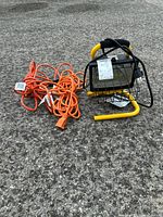 Portable work light with yellow metal stand and protective cage next to two orange extension cords laid on asphalt surface.
