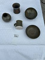 Photo showing four ceramic bowls, one small rounded vase, one small cup, and one signed mug arranged on white fabric outdoors.